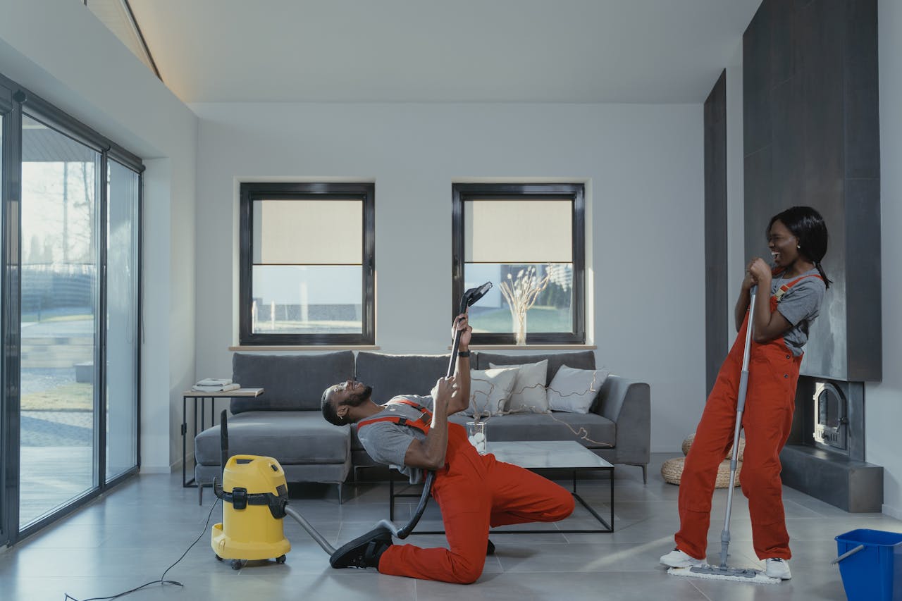 services-04 Fun and engaging cleaning session with a man and woman in overalls using cleaning tools in a modern living room.