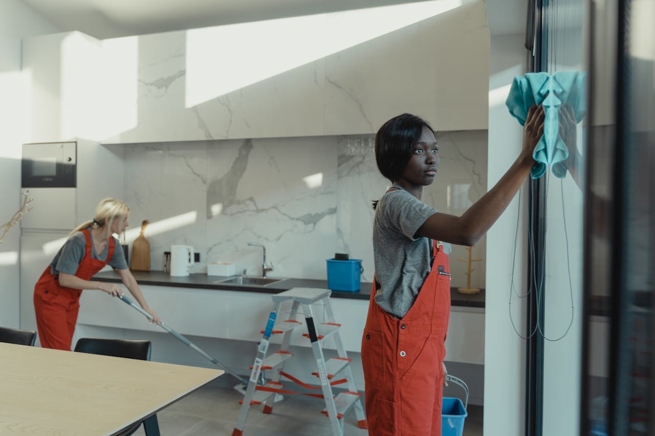 about-01 Two women cleaning a modern kitchen, wiping glass surfaces and mopping the floor.
